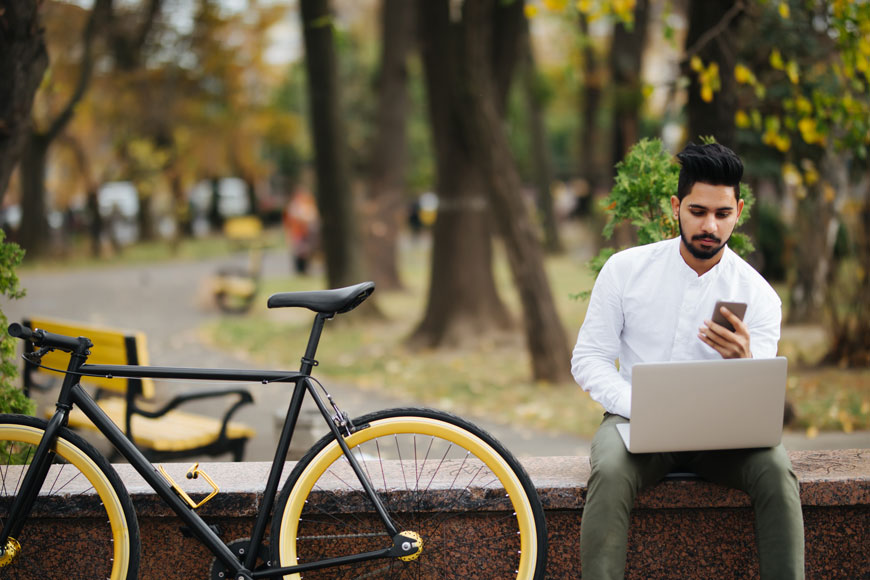 一个骑自行车的人坐在他的自行车旁边的公园里，用手机和笔记本电脑查看社交媒体上关于自行车的新闻。