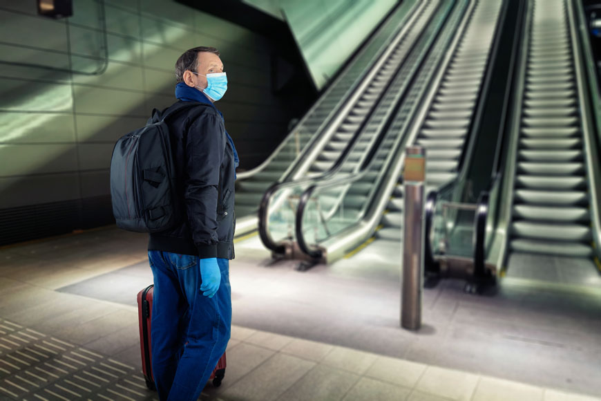 一个戴着蓝色纸外科口罩的男人站在前面有护柱的自动扶梯旁。