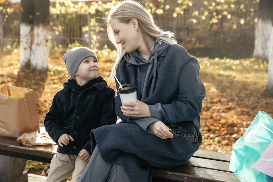 一个妈妈和孩子坐在一条靠背的公园长椅上，分享小吃，他们的包裹蔓延着。