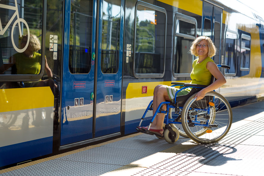 坐在轮椅上的女人等待在平台上登上公共汽车，该平台上有可访问的设计元素，例如两个高度的按钮，明亮的对比明确的信息和可检测的警告板