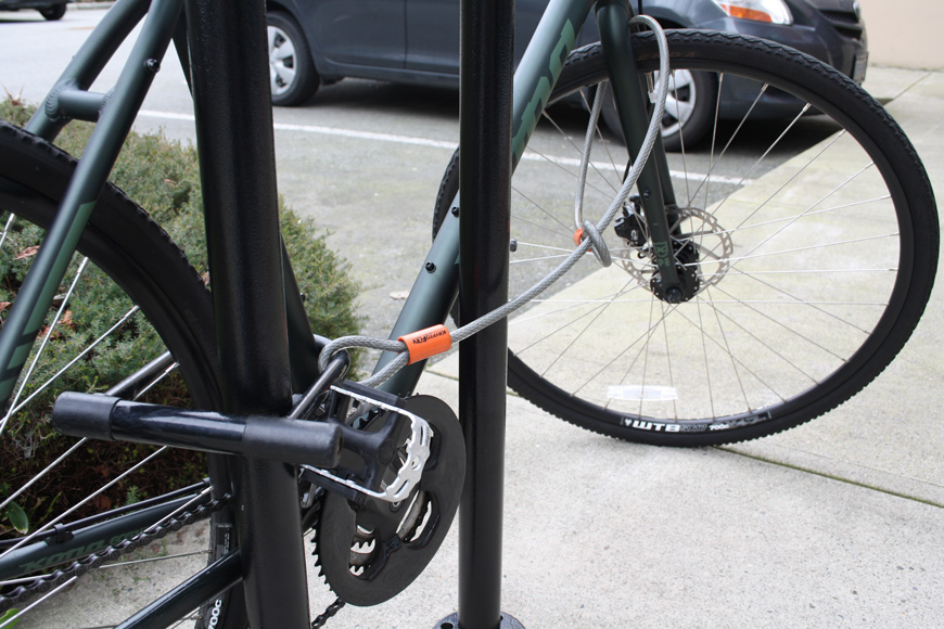 一辆深绿色的自行车被一个u型锁锁在后三角形，电缆连接到前轮