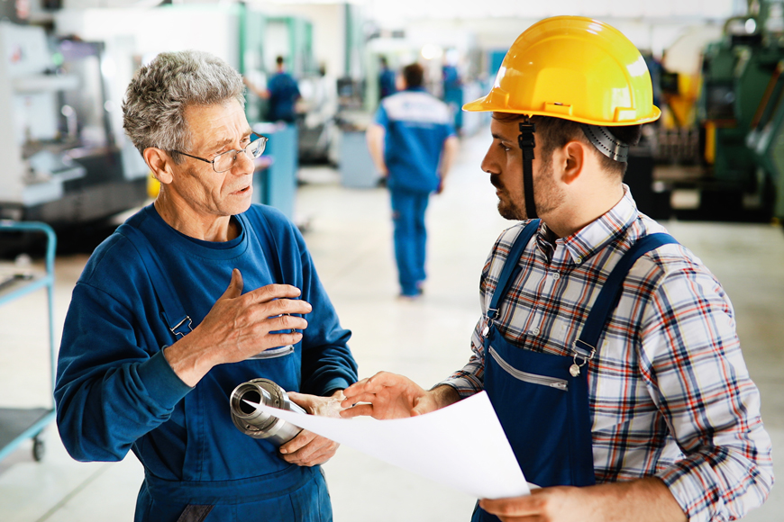 一位身穿蓝色制服的年长金属工人拿着一个零件，与一位安全帽管理员讨论数据