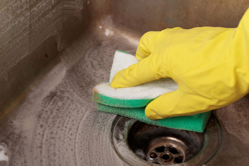 特写镜头，戴着黄色橡胶手套的手正在用温和的研磨剂清洗一个旧的不锈钢水槽