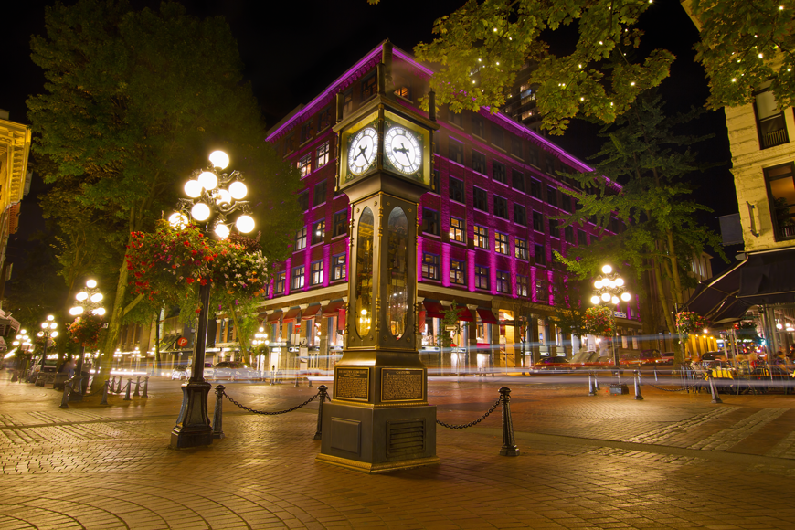 一个发光的夜景在鹅卵石街上有一个老式的蒸汽钟，后面有紫色的建筑物