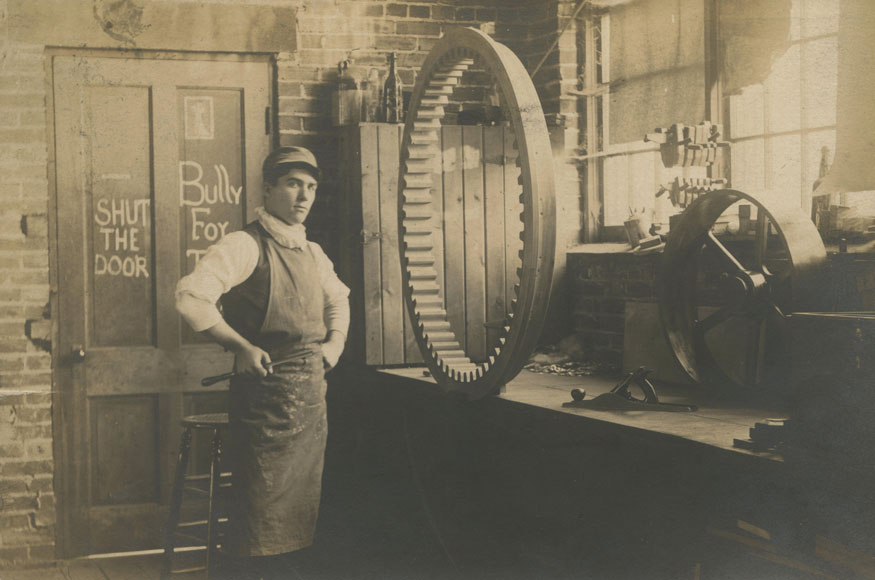 一张古色古香的明信片上的图片显示了一个年轻人站在一个巨大的木制齿轮前