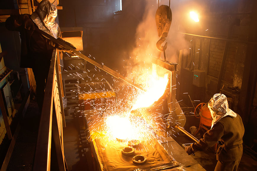 两名身穿银质隔热防护服的铸造厂工人用一个倾斜的钢包将金属倒入模具中