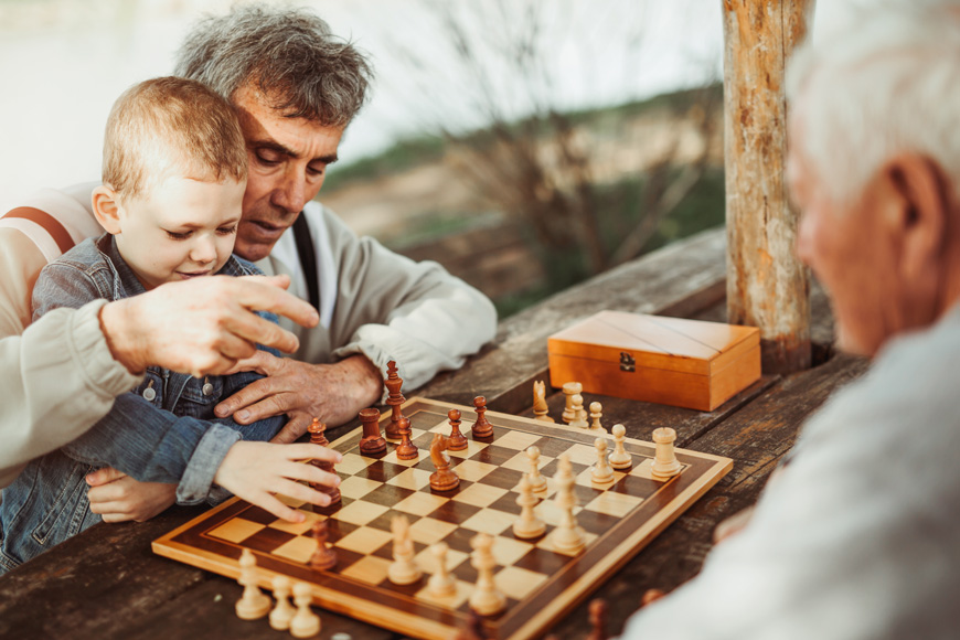 两个年长的男人在公园里下棋：一个将他的孙子握在膝盖上。