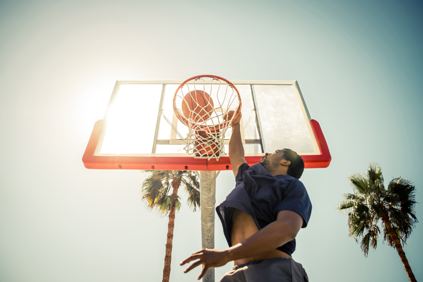 一名男子跳过相机上方，将篮球扣在阳光明媚的球场上，周围是棕榈树。