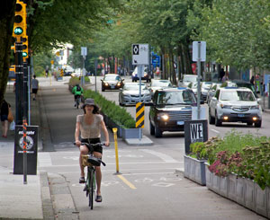 Two cyclists ride in opposite directions on a protected bike lane beside a busy downtown street, Vancouver BC