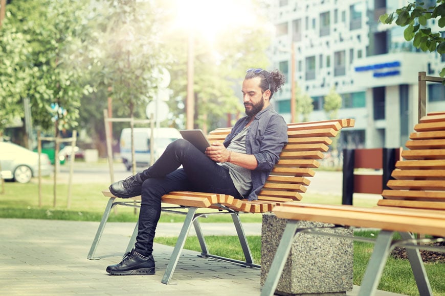 一名男子坐在城市公园的长椅上阅读平板电脑，身后是办公大楼和树木